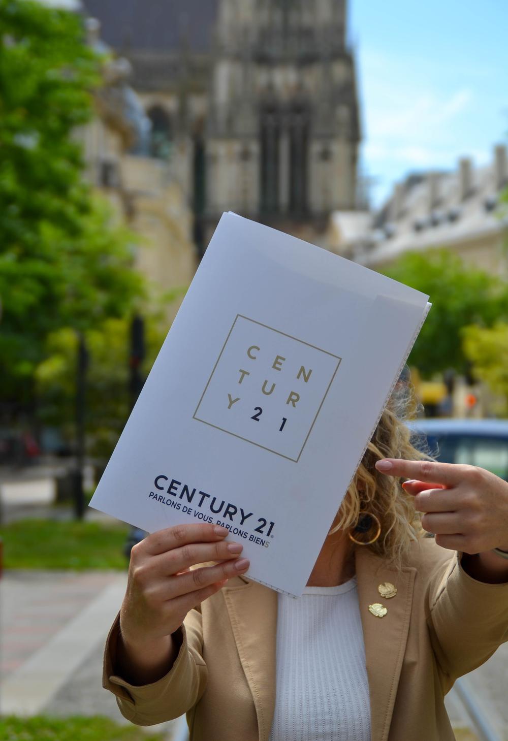 Une conseillère CENTURY 21 Martinot Immobilier montrant une pochette blanche Century 21 devant la Cathédrale de REIMS
